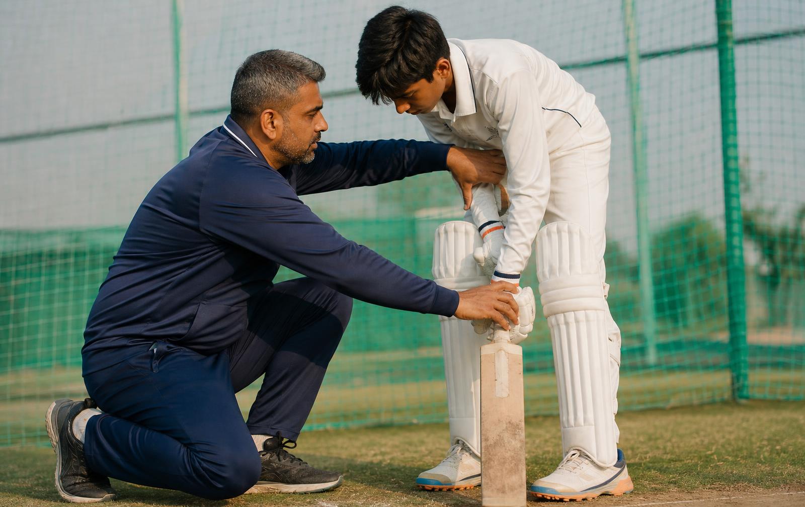 Coach kneeling beside a young batsman to correct his grip and stance